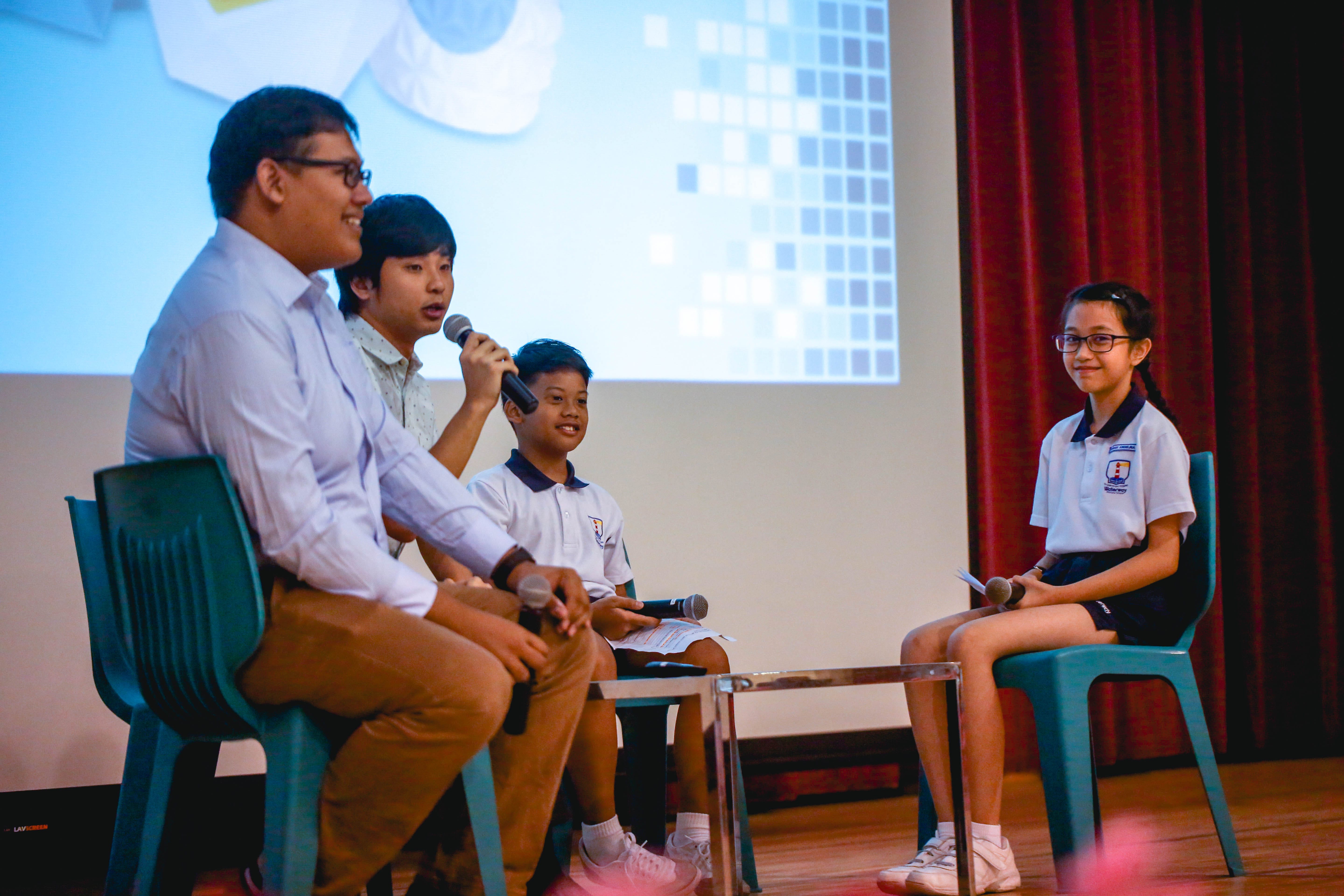 Four people sit on a stage with microphones, including two adults and two uniformed students, facing each other in discussion.